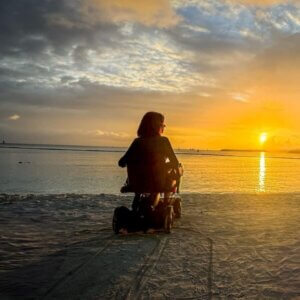A person in a motorized wheelchair, having arrived in a wheelchair van, faces the ocean, watching the sunset over the water from a sandy beach.