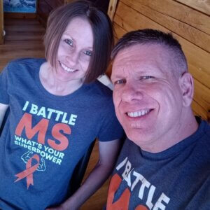 Two people smiling indoors, both wearing matching T-shirts that read I BATTLE MS WHATS YOUR SUPERPOWER with an orange ribbon graphic, pose proudly beside their accessible wheelchair van.