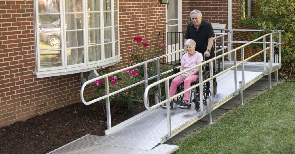 An elderly woman in a wheelchair is being pushed down an outdoor metal ramp by an older man next to a brick house with flowers nearby.