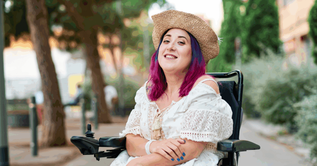 Woman with purple hair and a straw hat sits in a wheelchair outdoors, wearing a white dress and smiling. Trees and buildings are in the background.
