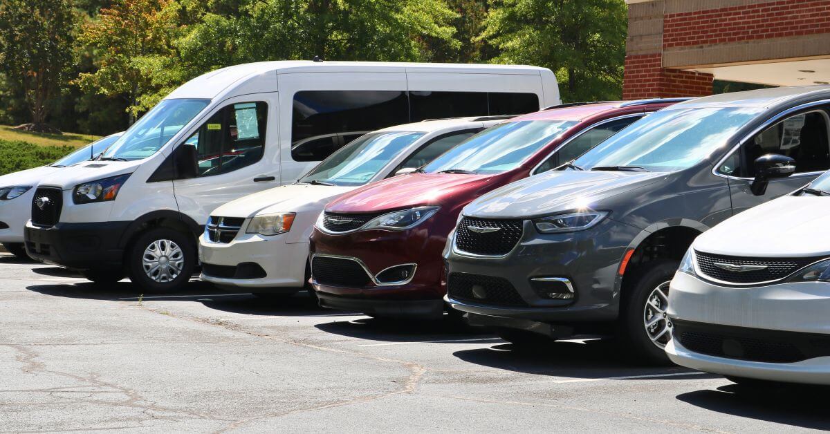 Several wheelchair minivans and a white passenger van are parked side by side in a parking lot near a brick building and trees.