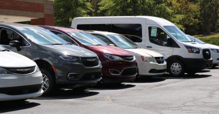 A row of parked minivans and a white passenger van in a parking lot on a sunny day.