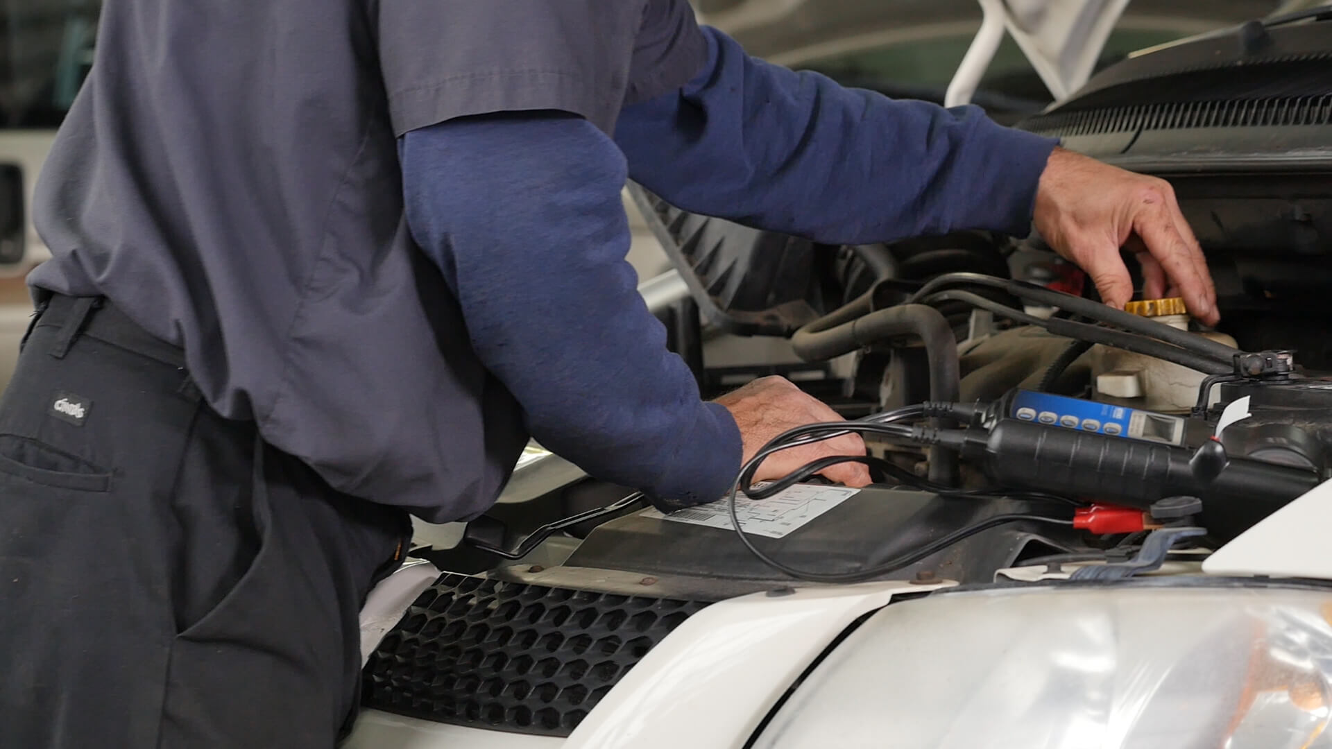 A person in a work uniform is checking under a preowned wheelchair van’s hood, using diagnostic tools and cables connected to the vehicle’s battery.