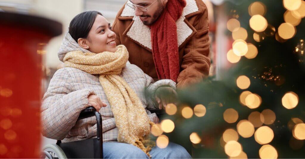 A woman in a wheelchair, bundled in winter clothing and a scarf, smiles at a man standing behind her near a decorated Christmas tree with lights.