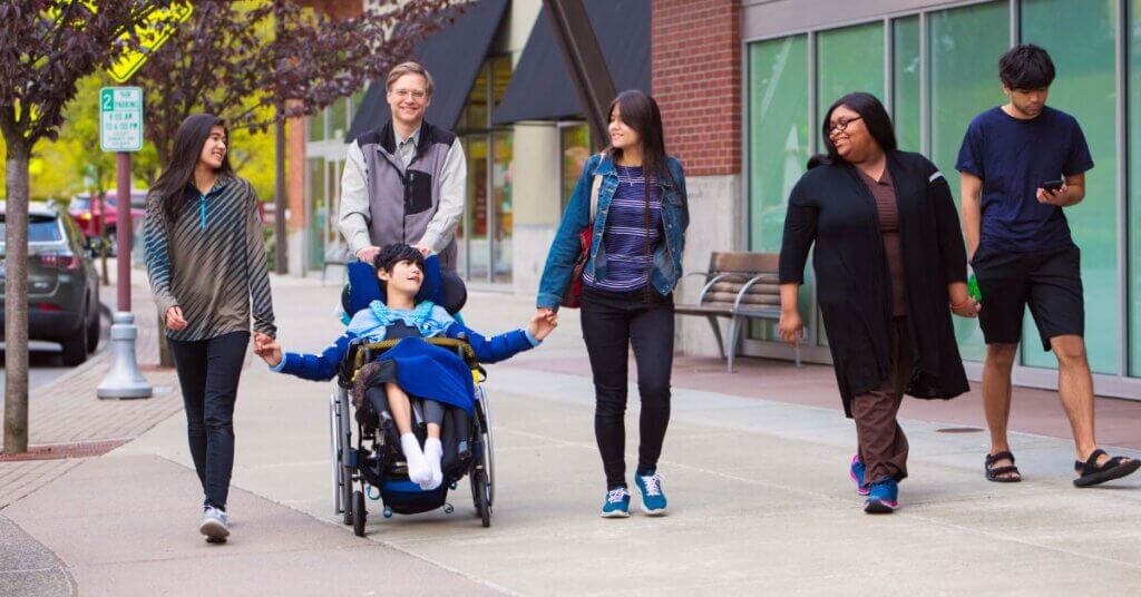 Six people, including a child in a wheelchair, walk together on a city sidewalk, smiling and interacting as vehicles pass by on a bright day.