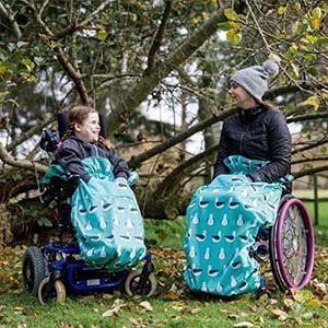 A child and an adult, each in wheelchairs, smile outdoors while wearing a BundleBean blanket