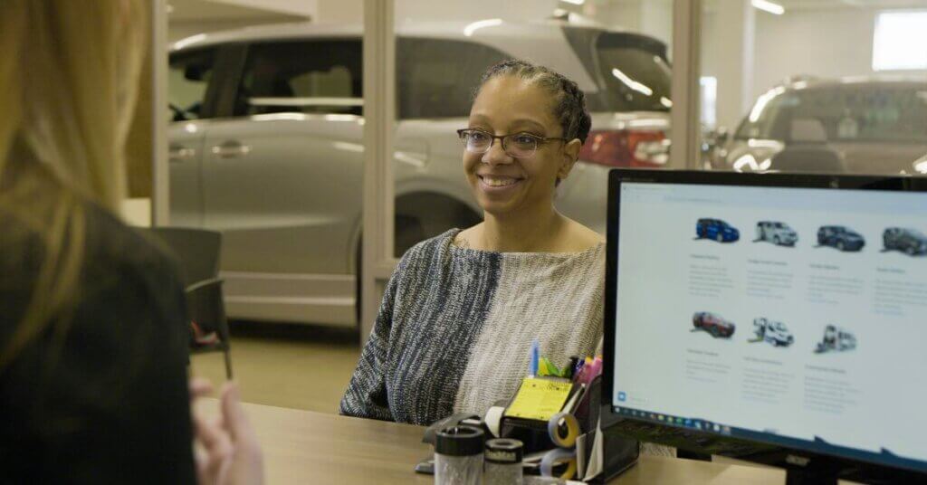 A woman smiles while sitting at a desk in a car dealership office. A salesperson faces her across the table with a computer displaying vehicle options.
