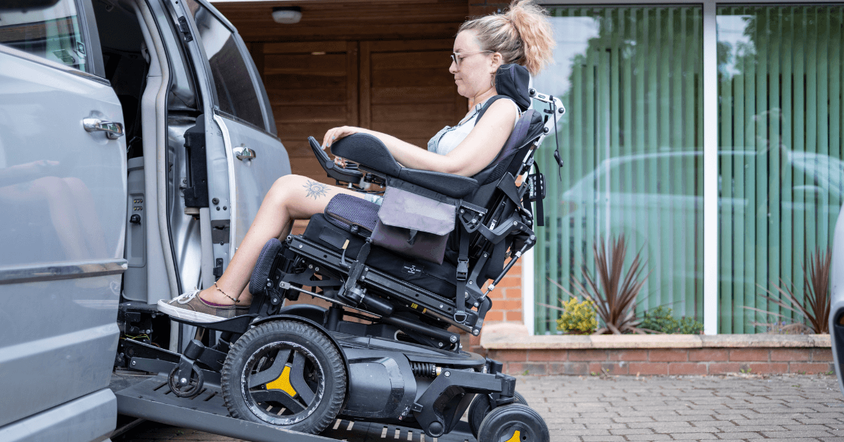 A woman in a motorized wheelchair uses a ramp to enter a van outside a building with large windows.