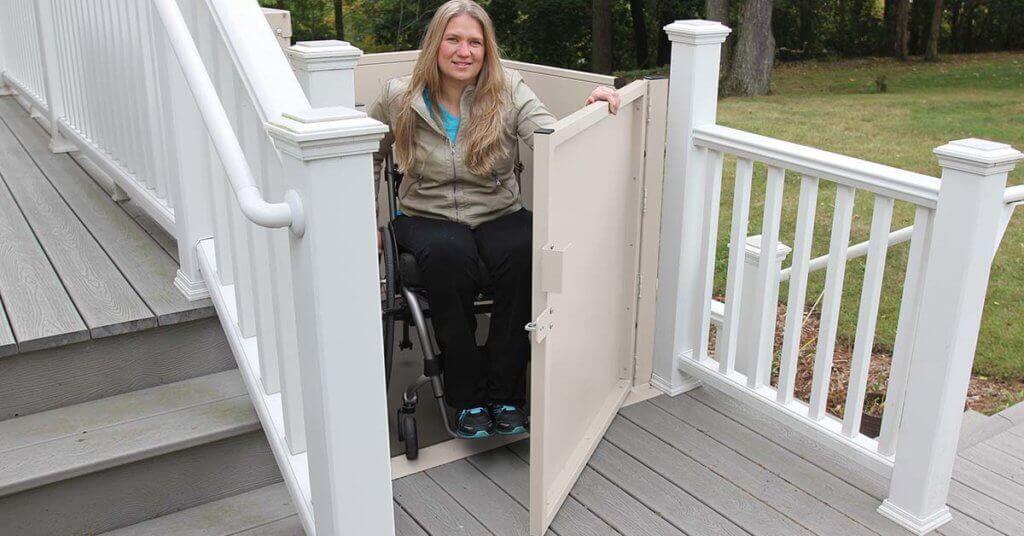 Una mujer en silla de ruedas utiliza una pequeña plataforma elevadora exterior para desplazarse entre los diferentes niveles de una terraza de madera rodeada de barandillas blancas.
