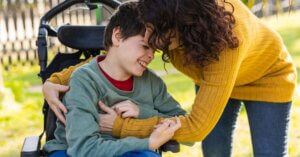 A woman in a yellow sweater hugs a smiling boy in a wheelchair outdoors on a sunny day, highlighting the importance of mobility solutions for caregivers.