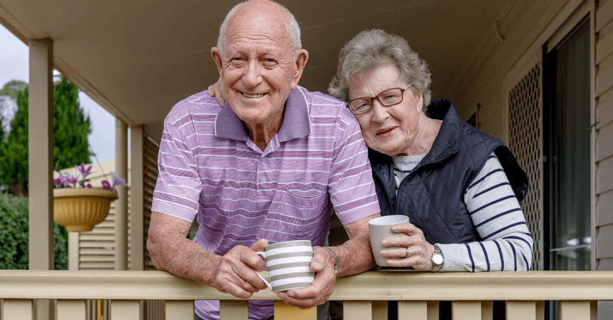 An elderly man and woman stand on a porch, smiling at the camera while holding coffee mugs. The man wears a pink striped shirt; the woman wears glasses, a striped top, and a dark vest.