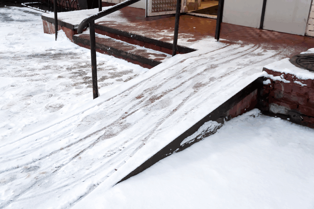 A snow-covered wheelchair ramp leads to a building entrance, with ice and snow visible on the surrounding ground and steps.