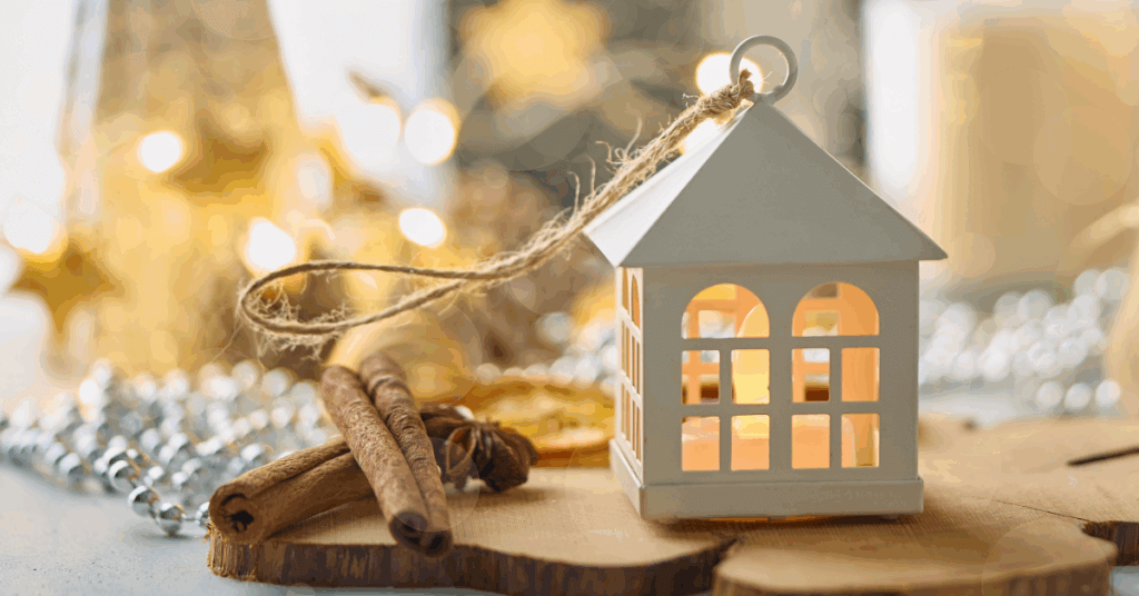 White lantern with a lit candle inside sits on a wooden surface next to cinnamon sticks and dried orange slices, surrounded by festive lights and ornaments.