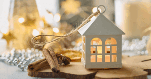 White lantern with a lit candle inside sits on a wooden surface next to cinnamon sticks and dried orange slices, surrounded by festive lights and ornaments.