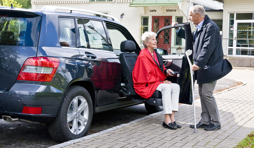 woman getting out of a blue vehicle using a turning seat with man holding door for her