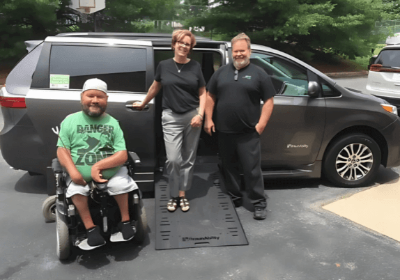 Three people by a parked van with an extended wheelchair ramp; one person is seated in a motorized wheelchair outside the van.