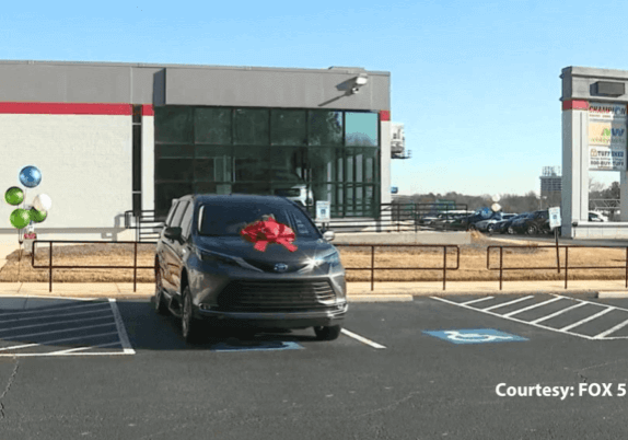 A minivan with a red bow is parked in a space near a building with large windows. Balloons are tied to a sign nearby.