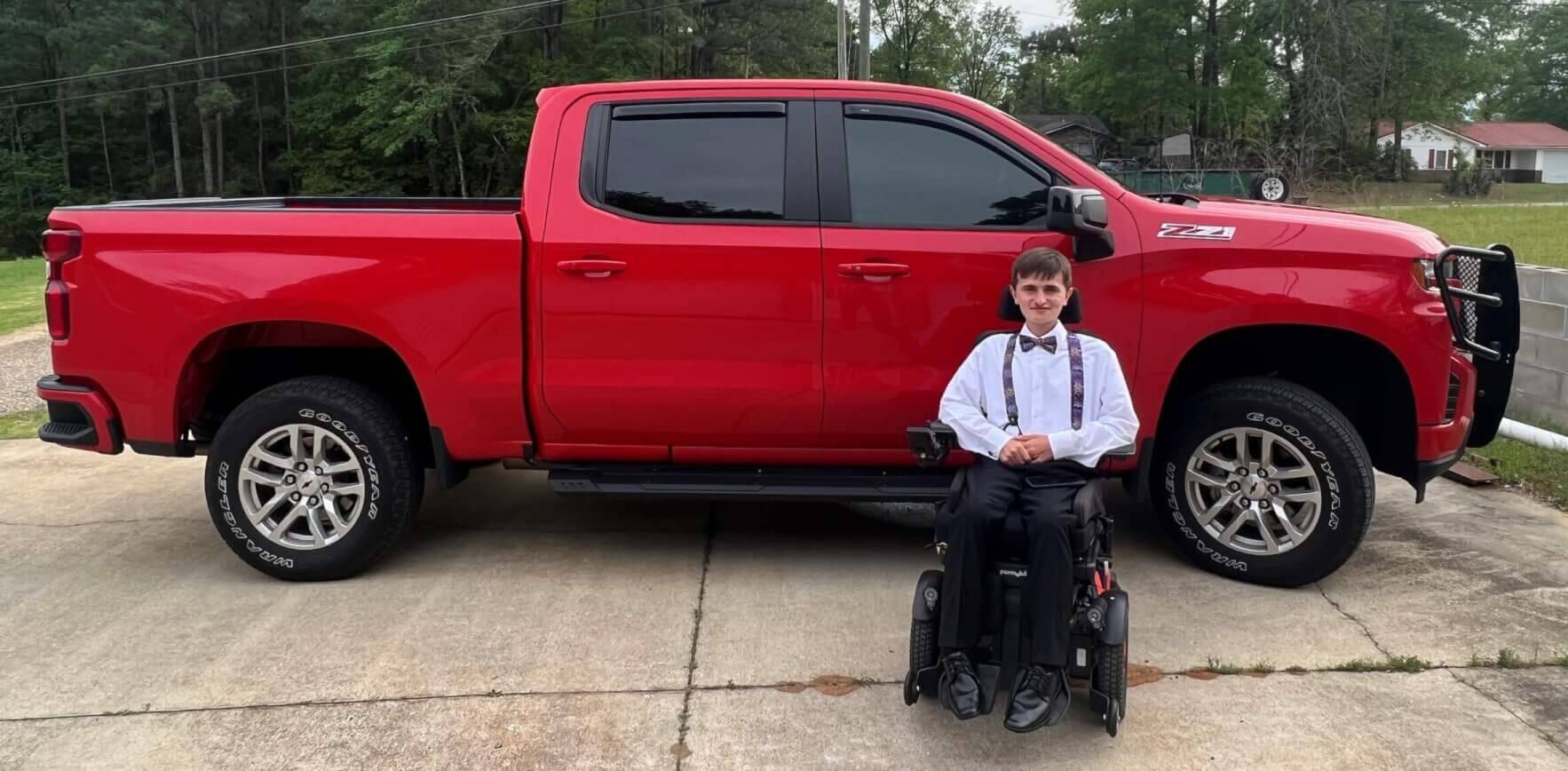 Estudiante de último año de secundaria, vestido con ropa formal, se sienta en su silla de ruedas frente a un Chevy Silverado 2022 de color rojo brillante