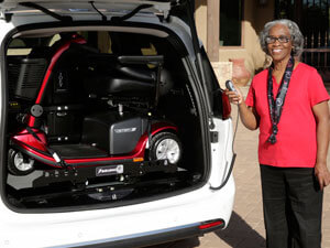 Woman standing holding control for scooter lift shown in back of vehicle.